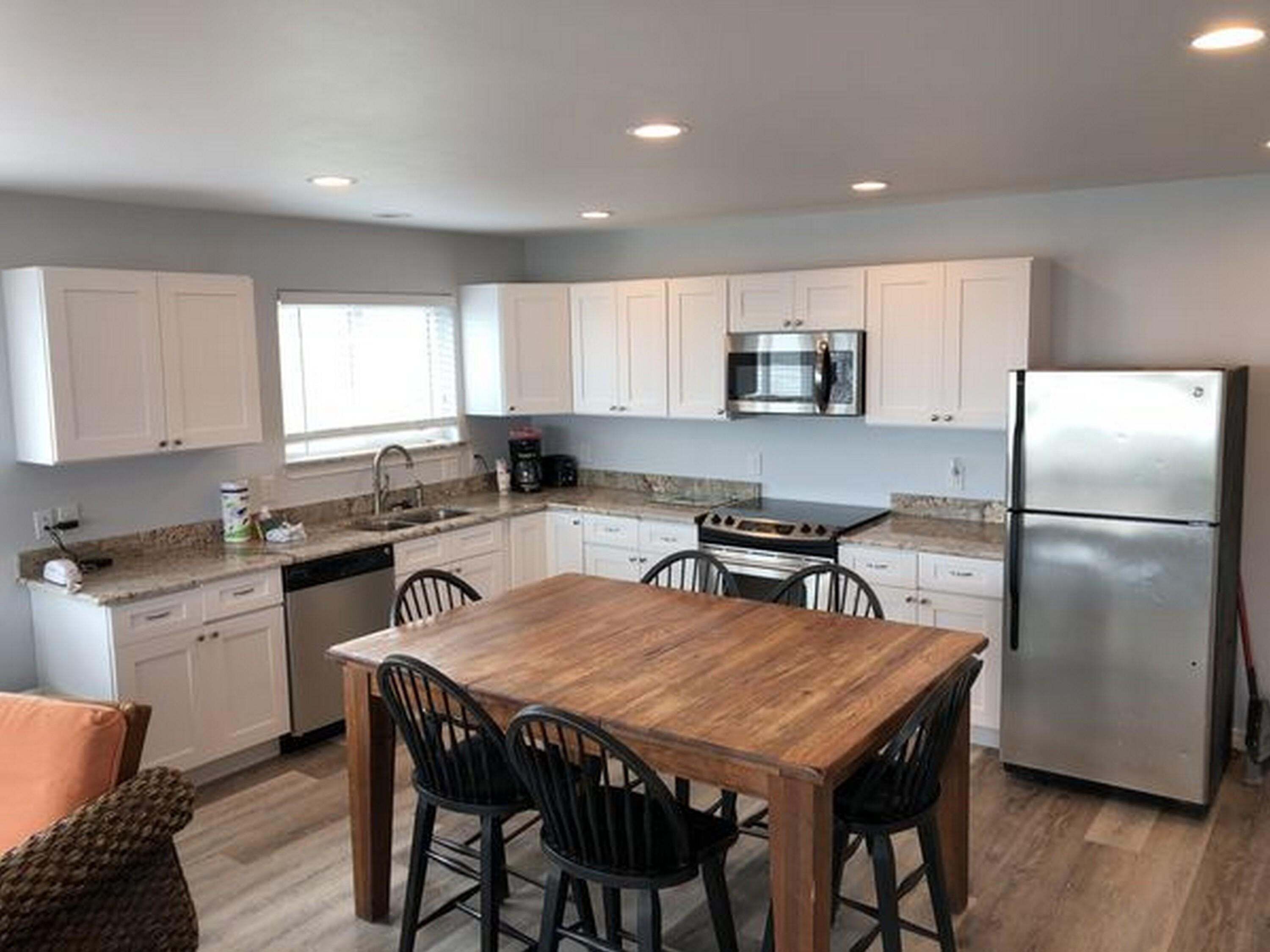 A modern kitchen with white cabinets, a stainless steel refrigerator, a built-in oven, and a wooden dining table with four chairs.