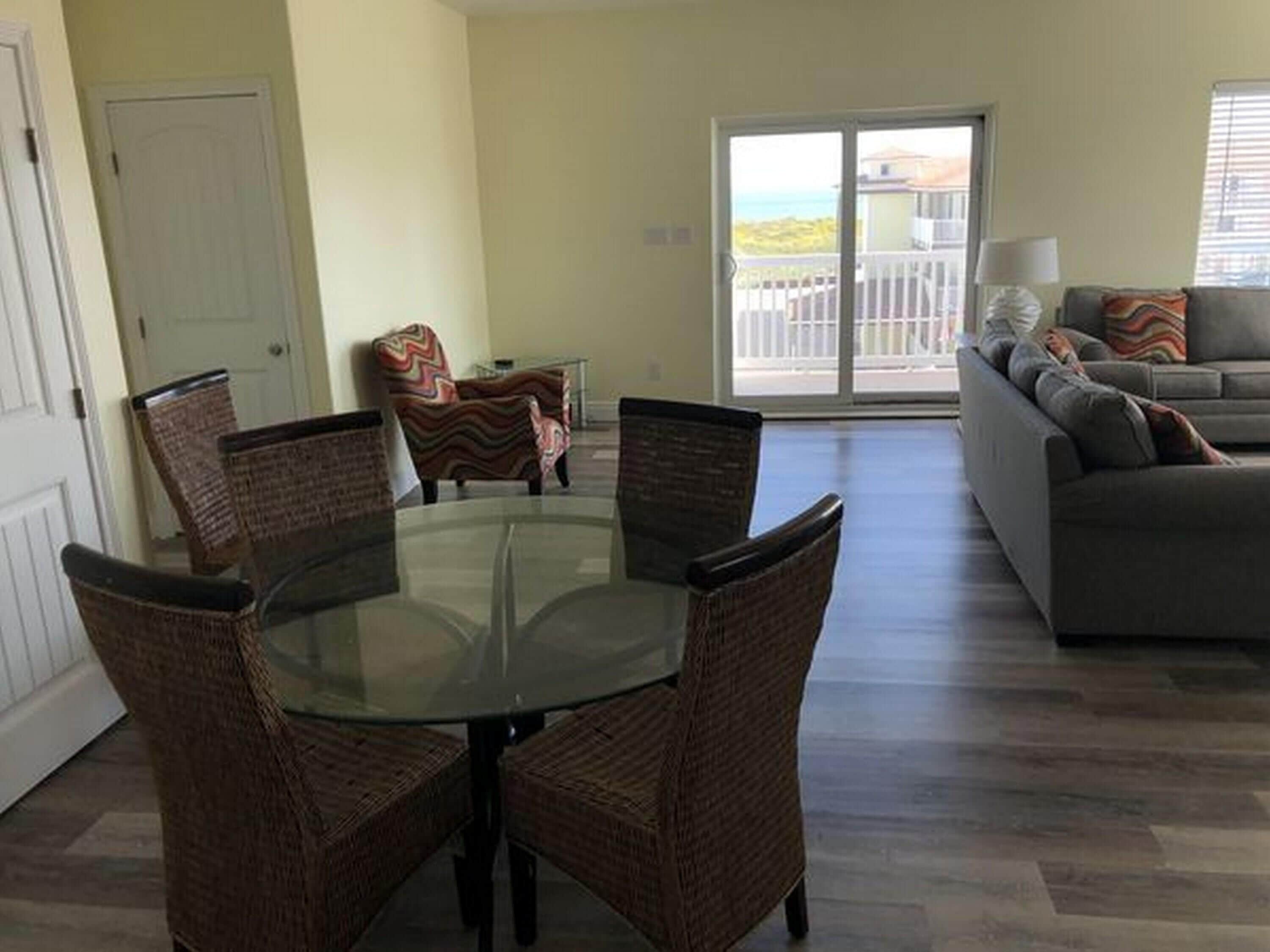A dining area with a glass table and wicker chairs, a sofa, and a view of the ocean through a sliding door.