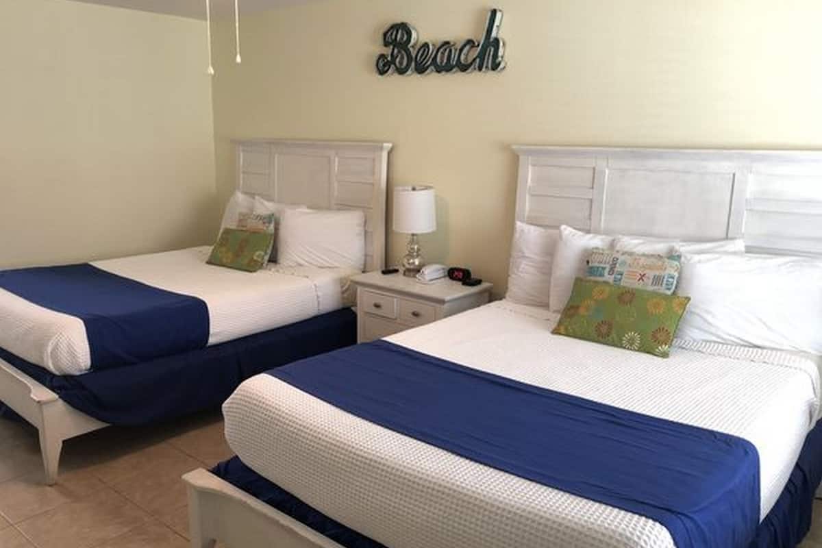 Two twin beds with white headboards and blue bedding, a bedside table with a lamp, and a wall sign that reads "Beach".