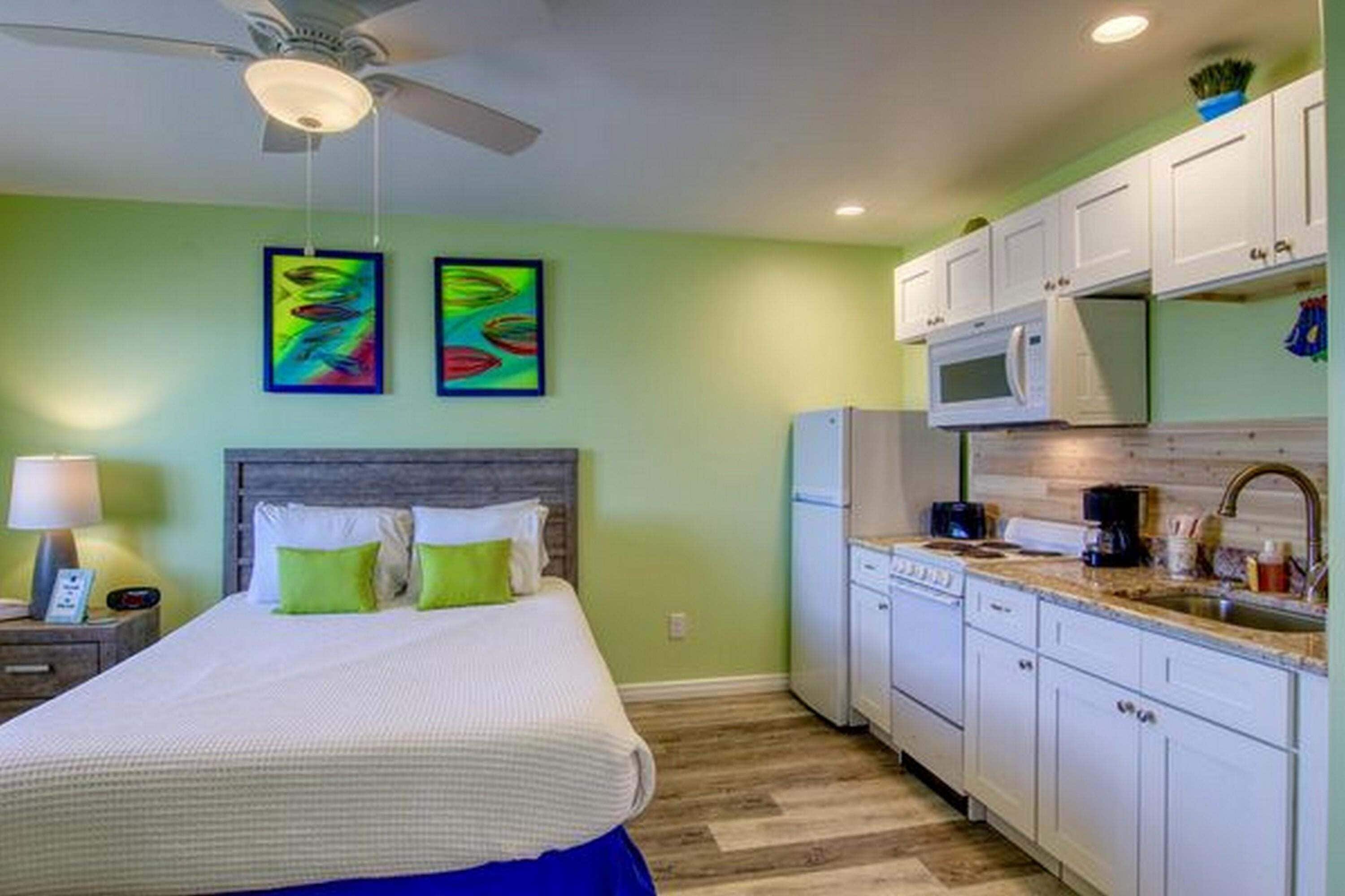 Quarto com cama, paredes verdes e ventilador de teto. Ao lado, cozinha com armários brancos, micro-ondas e pia.