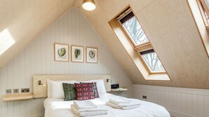 Ein Schlafzimmer mit schrägen Wänden, einem Oberlicht, einem Bett mit weißer Bettwäsche und drei gerahmten Pflanzenbildern an der Wand.