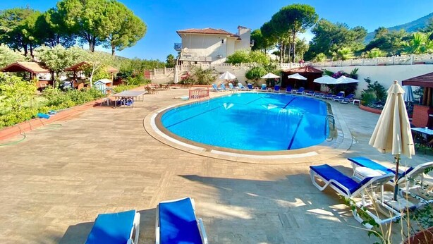 Une piscine entourée de chaises longues et de parasols, le tout bordé de verdure, avec un bâtiment visible au loin.