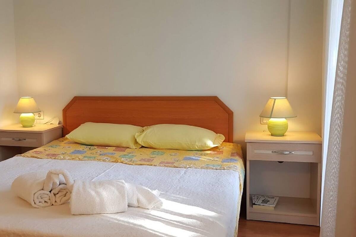 Un lit bien fait, recouvert d’une courtepointe, avec deux oreillers et des serviettes enroulées posées sur le dessus. Deux tables de chevet, chacune avec une lampe, se trouvent de chaque côté du lit.