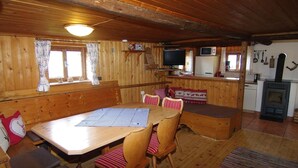 A cozy cabin interior with a wooden dining table, chairs, a bench, a kitchen area with a stove, and a television. - Quaint Mountain Hut in Hippach With Garden and Barbeque (Hippach)