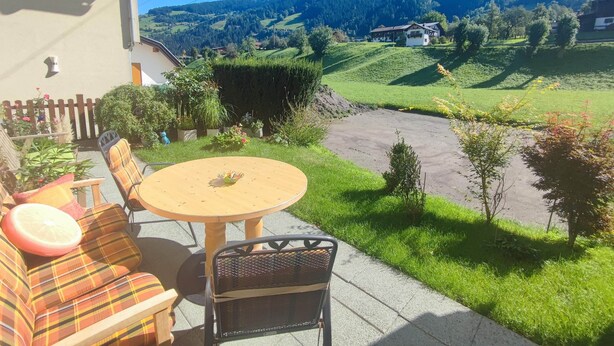 Eine Terrasse mit rundem Tisch und Stühlen, ein kleiner Garten und Blick auf eine Berglandschaft.