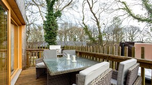 Eine Terrasse im Freien mit Korbmöbeln, einem Tisch mit Glasplatte und Blick auf Bäume sowie einen Holzzaun.