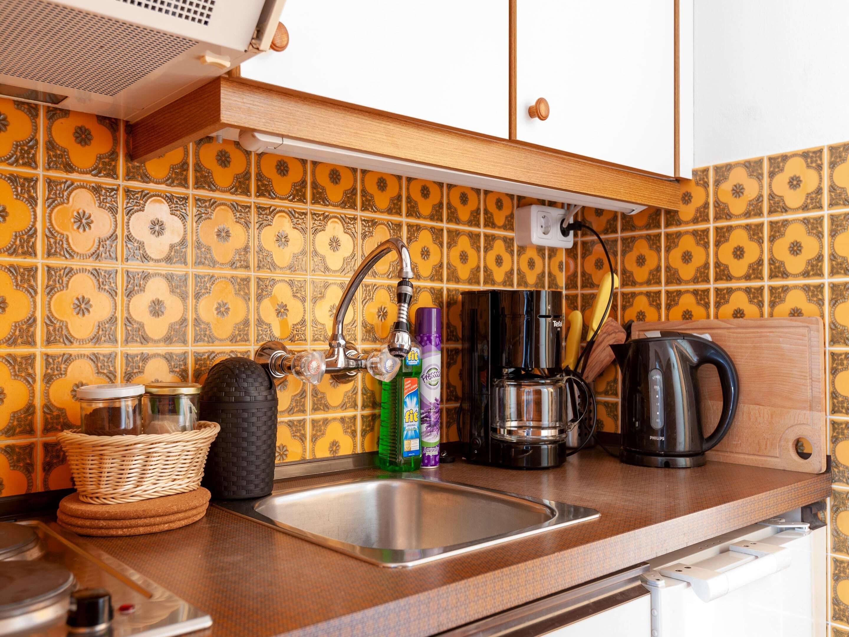 Uma cozinha com pia de aço inoxidável, cafeteira e torradeira. Há uma cesta e uma garrafa de detergente na bancada.