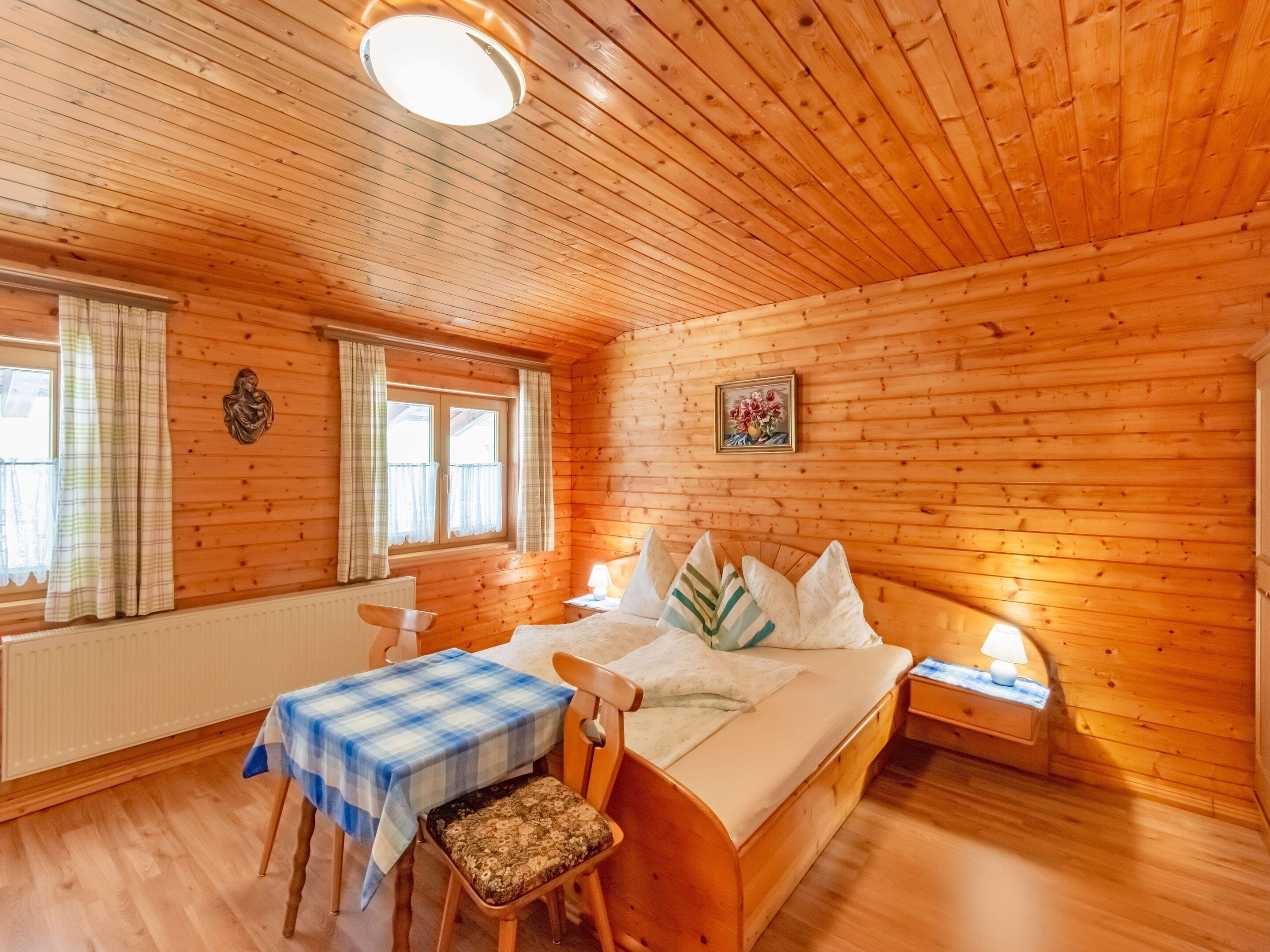 A cozy wooden room with a bed, a table with a checkered tablecloth, a chair, and a window with curtains.
