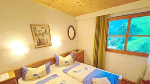 A neatly made bed with floral bedding, a bedside table with a lamp, a framed picture on the wall, and a window with a view of greenery. - Group Holiday Home in Hippach With Dreamy Views (Hippach)