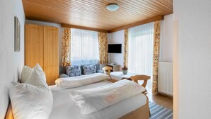 Quarto com teto de madeira, cama com roupa de cama branca, escrivaninha com cadeira e televisão na parede.