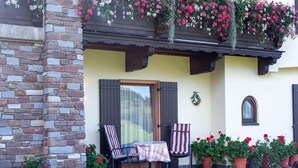 Um edifício de pedra com fachada amarela, varanda de madeira com cestos de flores pendurados e uma mesa posta com toalha quadriculada e cadeiras listradas.