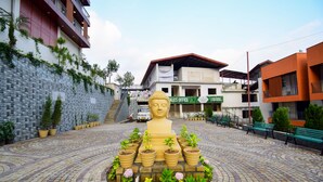 A Buddha statue surrounded by potted plants in a courtyard with a stone-paved area, buildings in the background, and a sign for a sales office. - Cliffton Valley by Dumnu Homes (Shimla)