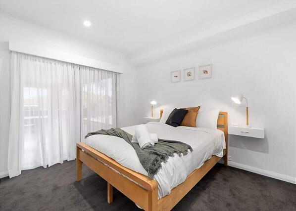 Un dormitorio moderno con cama de madera, paredes blancas y un ventanal con cortinas de gasa.