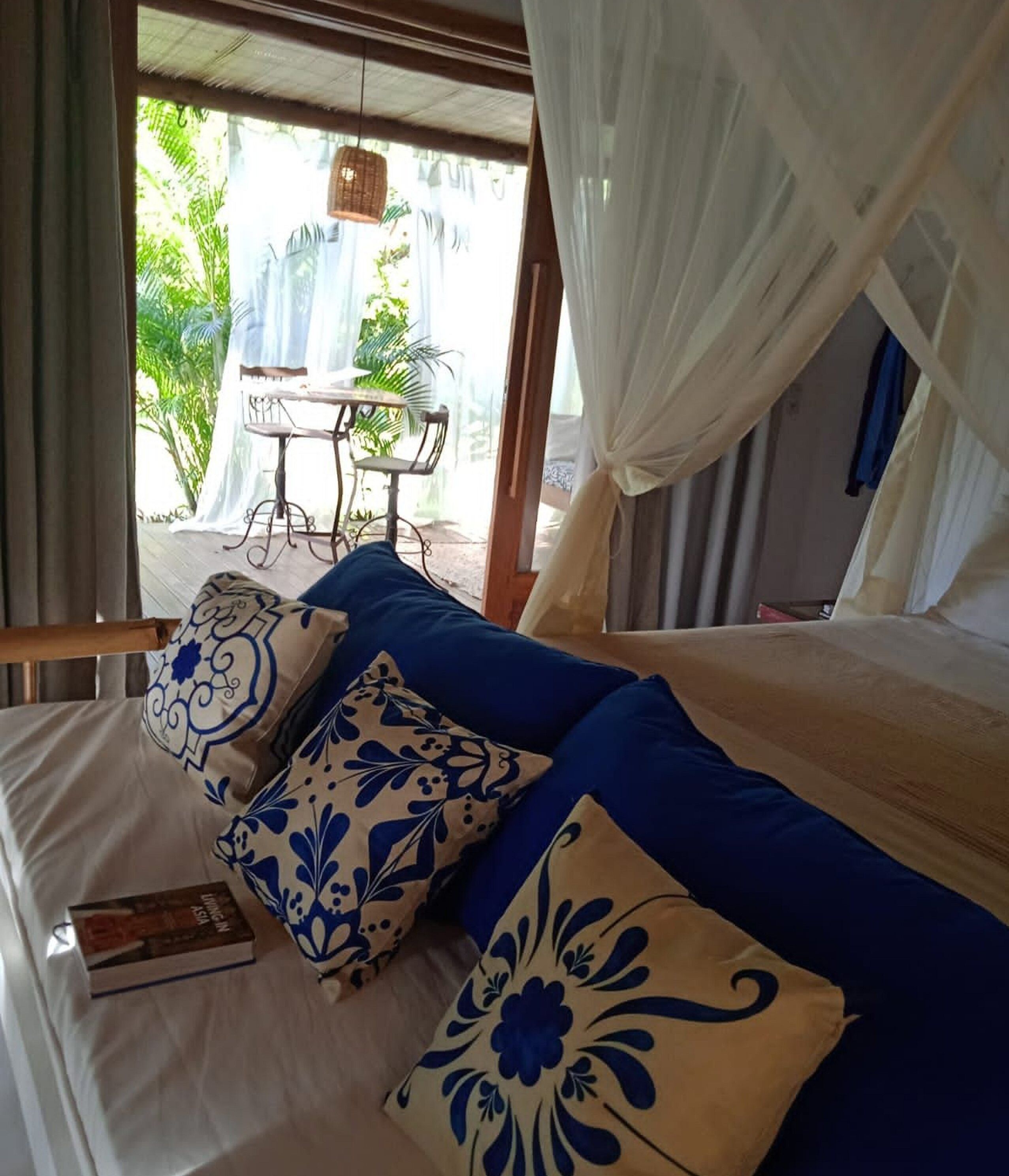 Ein Bett mit blau-weißen Kissen, ein Buch darauf und Blick auf einen Außenbereich mit Tisch und Stühlen.