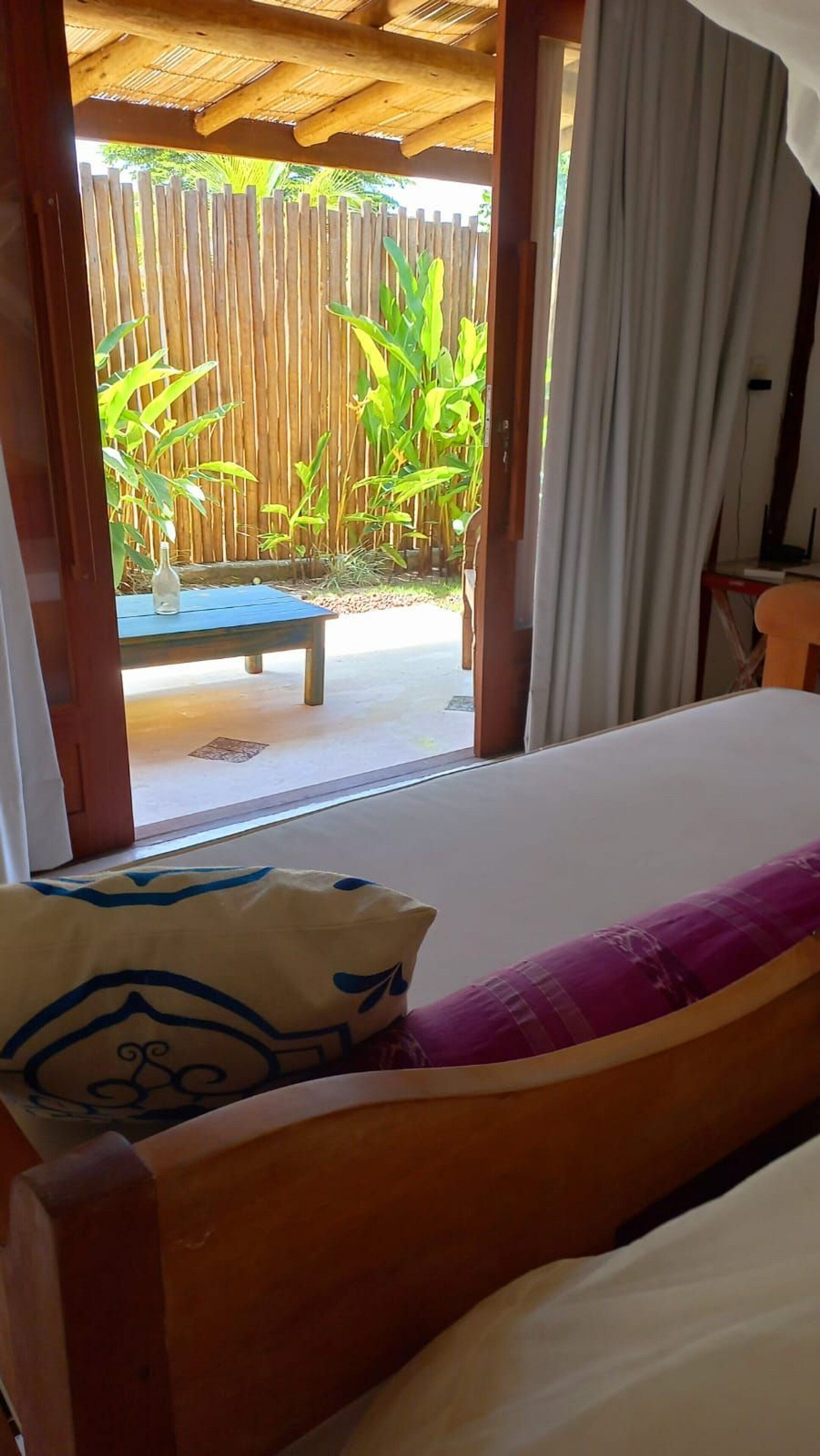 A bedroom with a bed, a wooden headboard, a window with curtains, and a view of a patio with a table and plants.