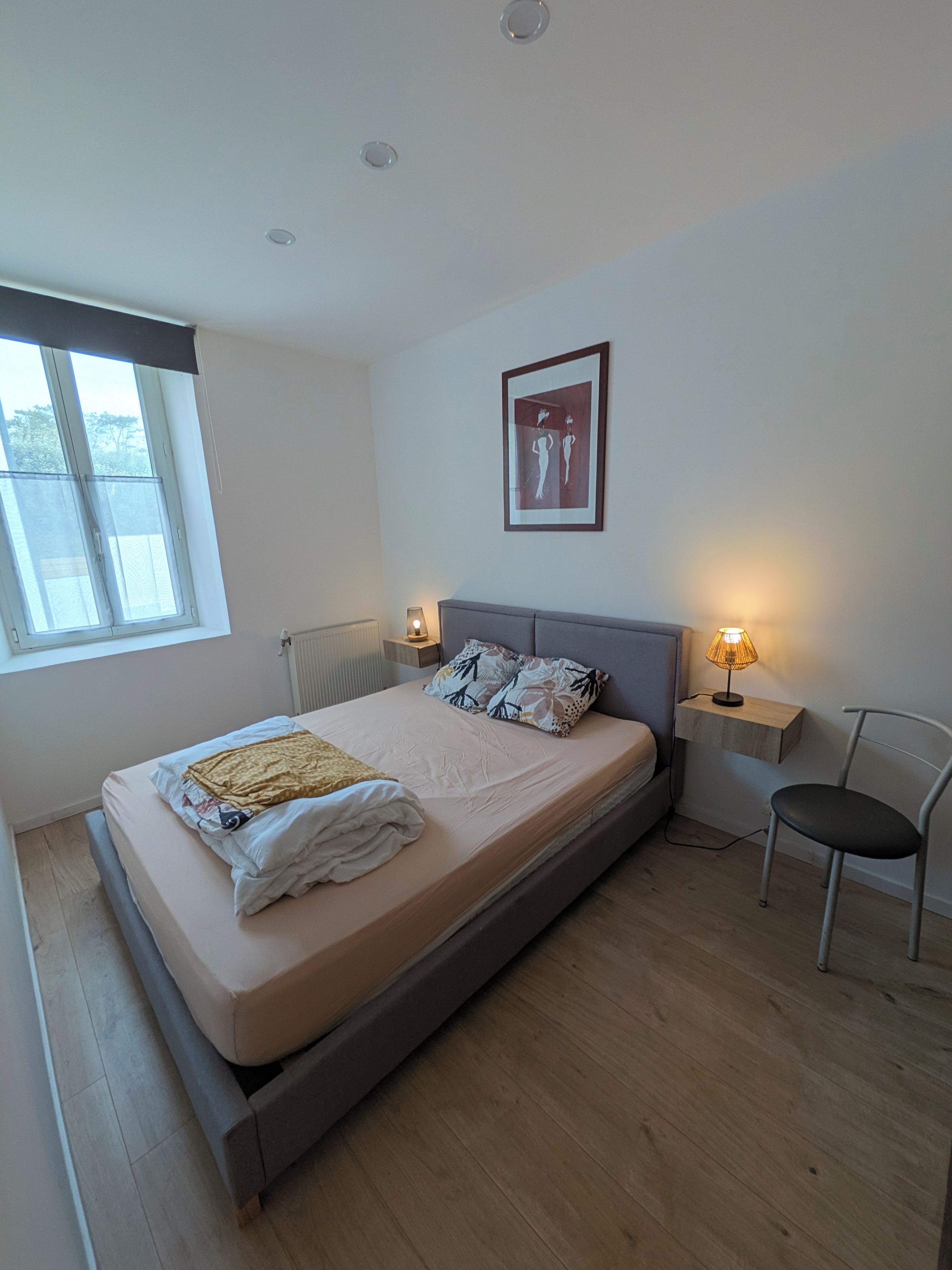 Quarto com cama, abajures, uma cadeira, janela e quadro pendurado na parede.