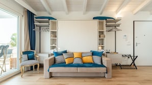 A modern living room with a sofa, a chair, and a desk. There is a large window with curtains, a ceiling with exposed beams, and a door with a handle. - Le Residenze di Carlà (Cagliari)