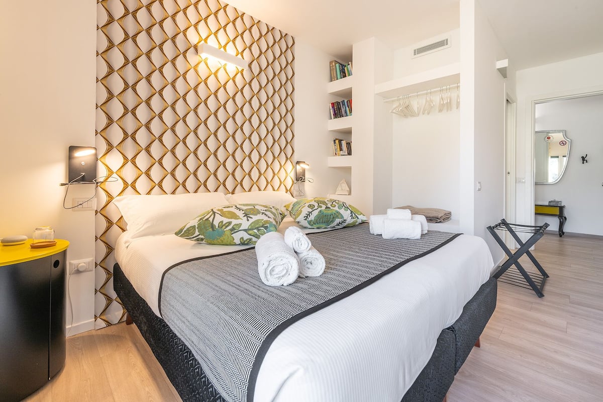 Un dormitorio moderno con un cabecero estampado, una mesita de noche con una lámpara, una cama con toallas enrolladas y un armario con espejo.