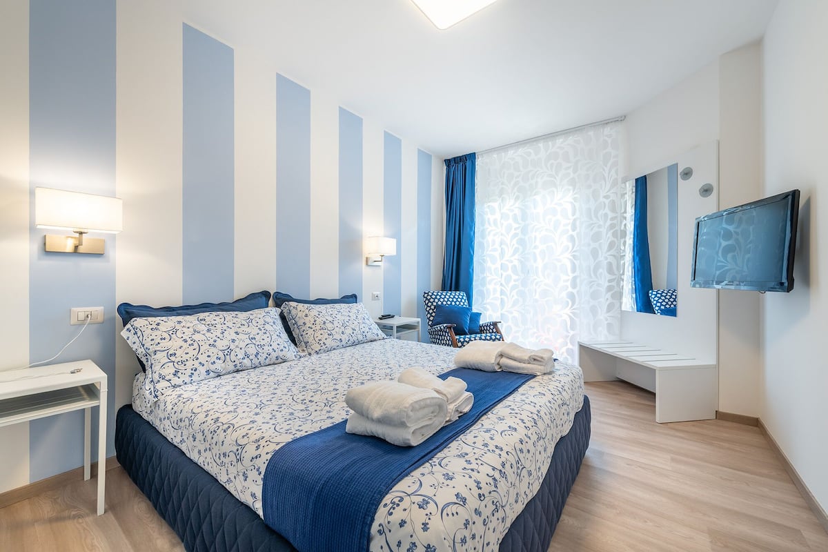 Habitación de hotel con una cama grande, un escritorio, una silla, un televisor y un ventanal con cortinas.