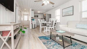 En moderne og lys stue med en hvid sofa, et sofabord og et spiseområde med hvide stole. Loftsventilator og fjernsyn er synlige.