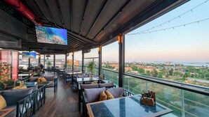 Un bar en la azotea con mesas al aire libre, grandes ventanales y vistas panorámicas de la ciudad.