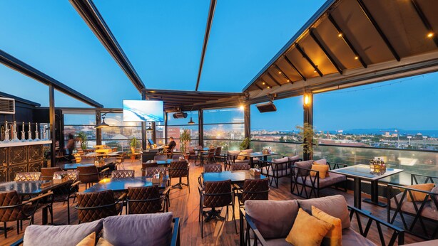 Un bar en la azotea con mesas al aire libre, ventanas amplias y vistas panorámicas de la ciudad.