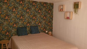 A bedroom with a patterned wallpaper, a bed with a quilted cover, two pillows, a small stool, and two wooden shelves with plants. - South-facing detached house with private pool (Puy-l'Évêque)