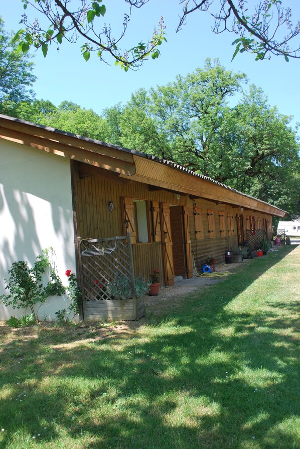 A row of wooden cabins with a covered entrance, a fenced area with potted plants, and a grassy lawn.