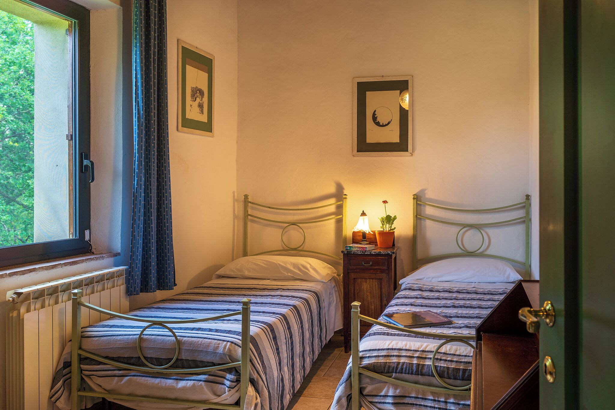 Slaapkamer met twee bedden, een raam, een radiator en een nachtkastje met een lamp en een plant.