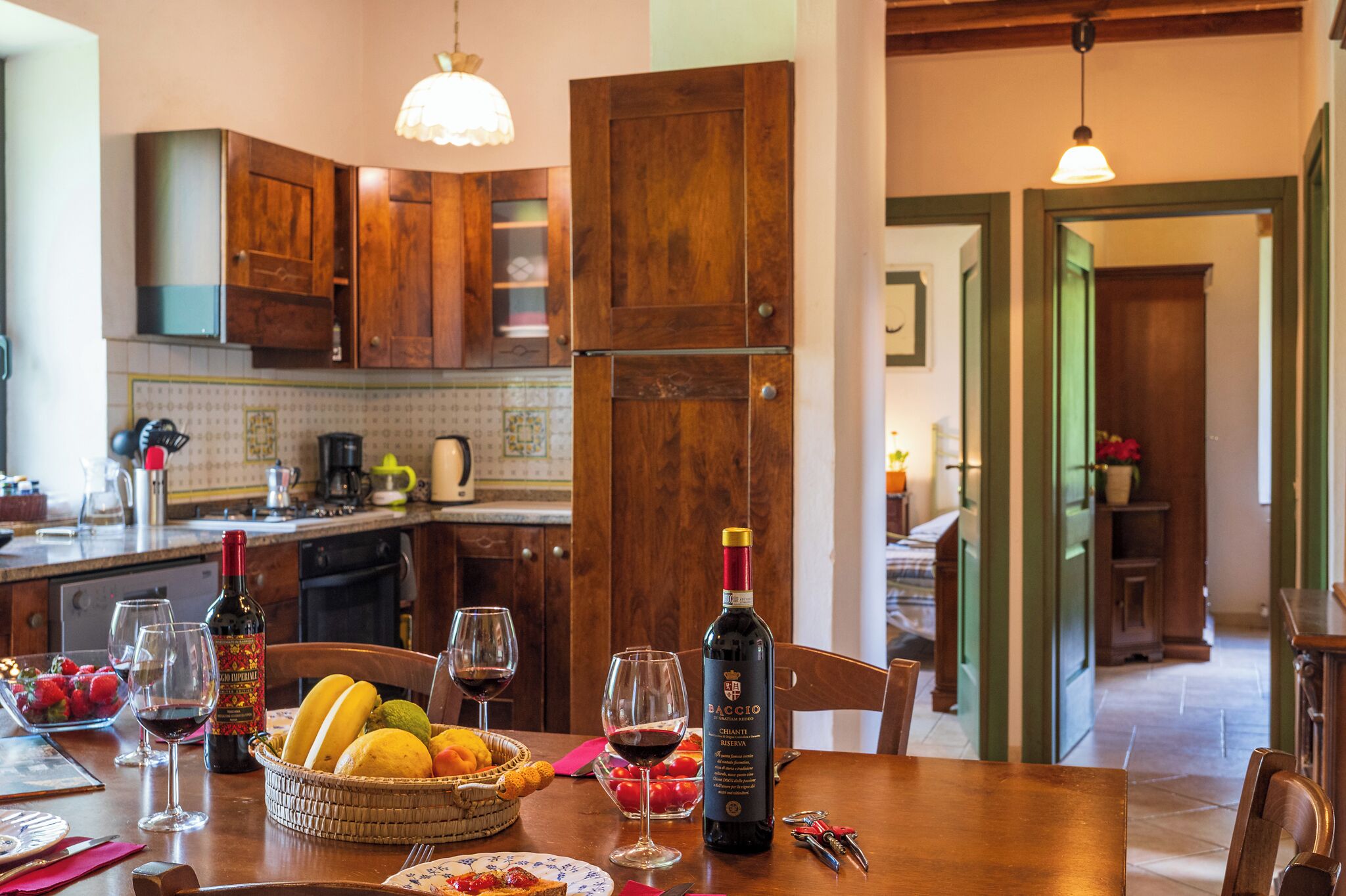 Une cuisine avec des meubles de rangement en bois, une table à manger dressée avec des verres à vin, une bouteille de vin et un panier de fruits.