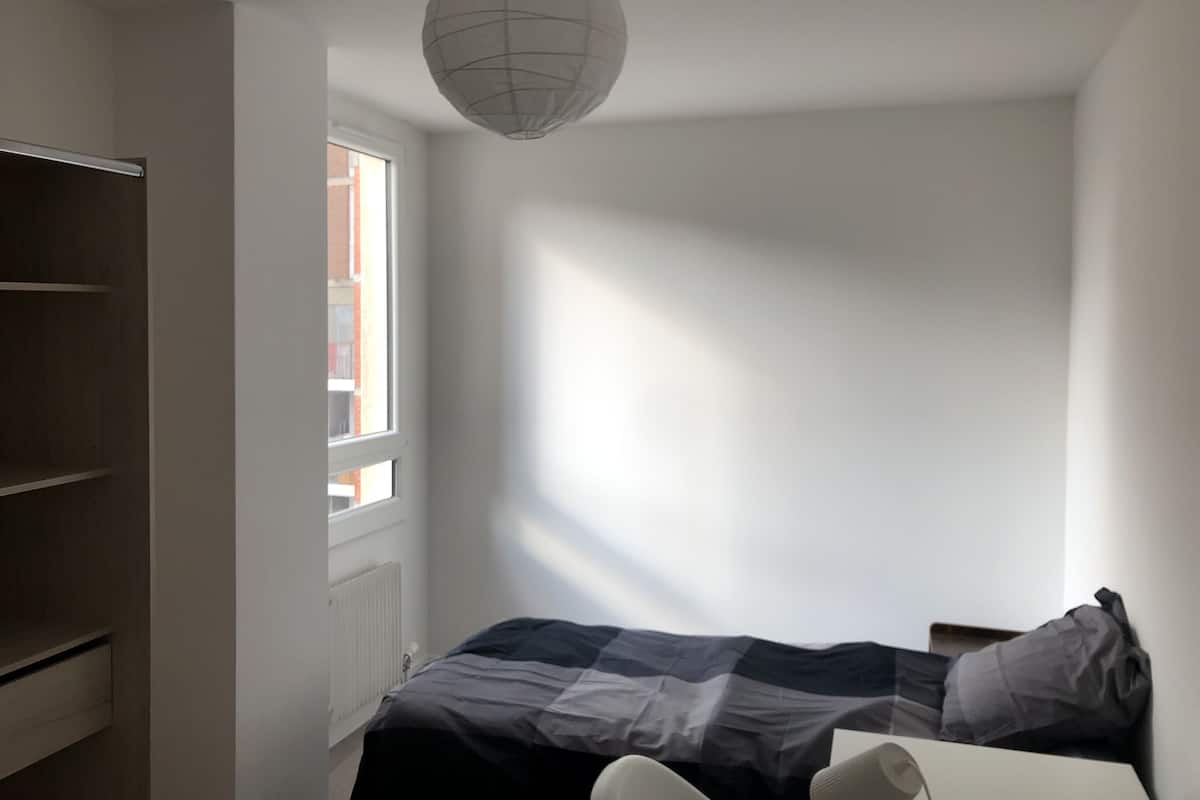 A bedroom with a bed, a radiator, a window, and a hanging light fixture.