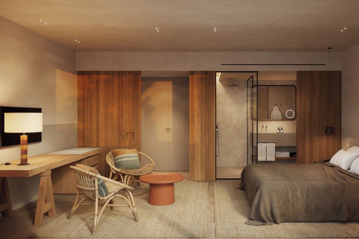 A modern hotel room with a bed, a desk, a chair, a small table, and a bathroom visible through an open door.