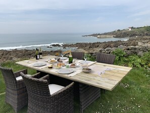 An outdoor dining table set on a grassy area with wicker chairs, overlooking a rocky coastline and the sea. - A house with the ocean at the end of the garden (Plozévet)