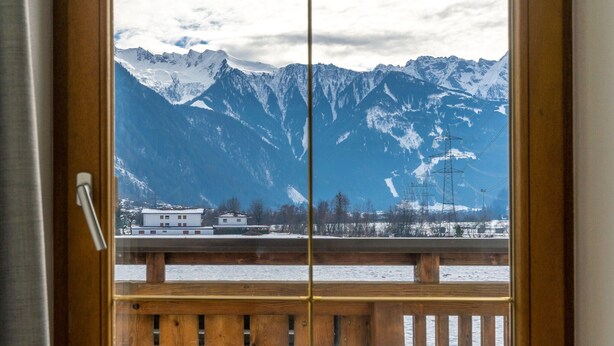 Vista a través de una ventana con baranda de madera, mostrando montañas nevadas y un edificio en primer plano.