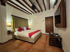 A hotel room with a bed, bedside lamps, a wooden headboard, a ceiling fan, a wooden door, and a small table with a vase. - VAARSA (Alibag)