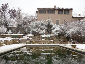 Ein mit Schnee bedeckter Garten mit einer Steinmauer und einem Pool, der die umliegenden Bäume spiegelt.