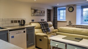 A compact kitchen with a dishwasher, a dining table with a notebook, a sofa, and a window with a clock. - 1 bedroom accommodation in Cemaes Bay (Cemaes Bay)