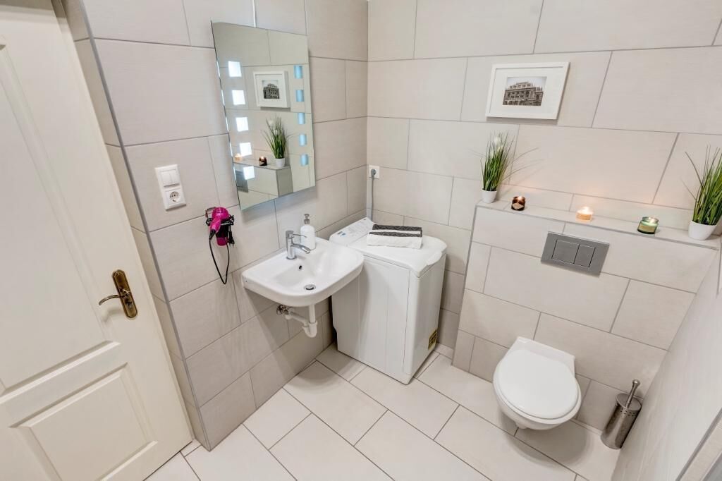 Une salle de bain moderne avec un lavabo, des toilettes et un miroir, le tout de couleur blanche. On y trouve également des plantes en pot et une petite étagère ornée d’objets décoratifs.