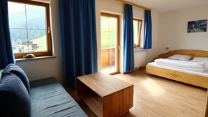 A room with a wooden bed, a sofa, a wooden table, and large windows with blue curtains. - Holiday Apartment in Stubai With Balcony (Neustift Im Stubaital)