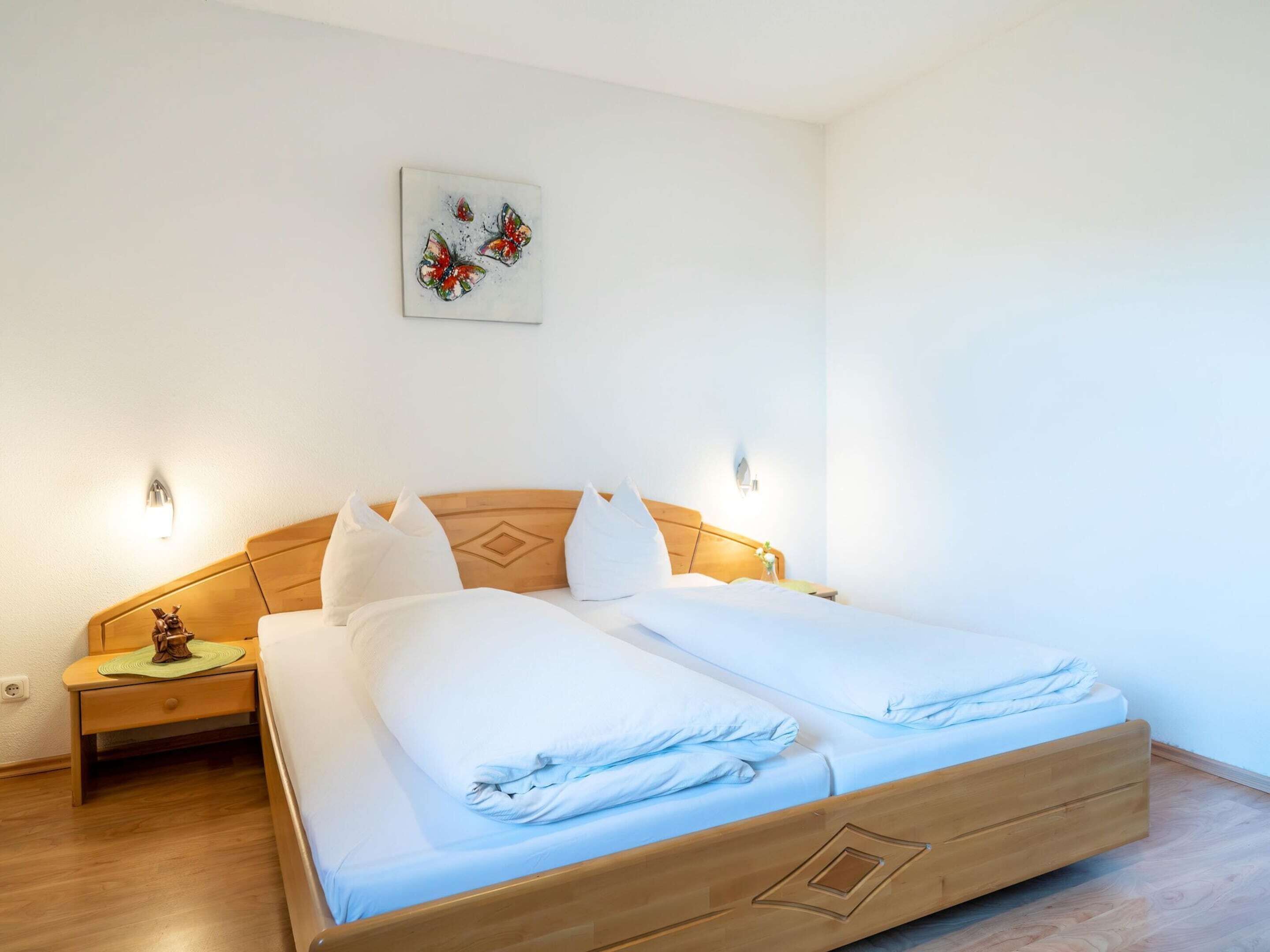 Ein Doppelbett mit Holz Kopfteil, weißer Bettwäsche, einem kleinen Nachttisch mit Lampe und einem an der Wand hängenden Kunstwerk.