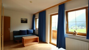 A room with a wooden floor, a sofa, a wooden coffee table, a bed, and large windows with blue curtains. - Holiday Apartment in Stubai With Balcony (Neustift Im Stubaital)