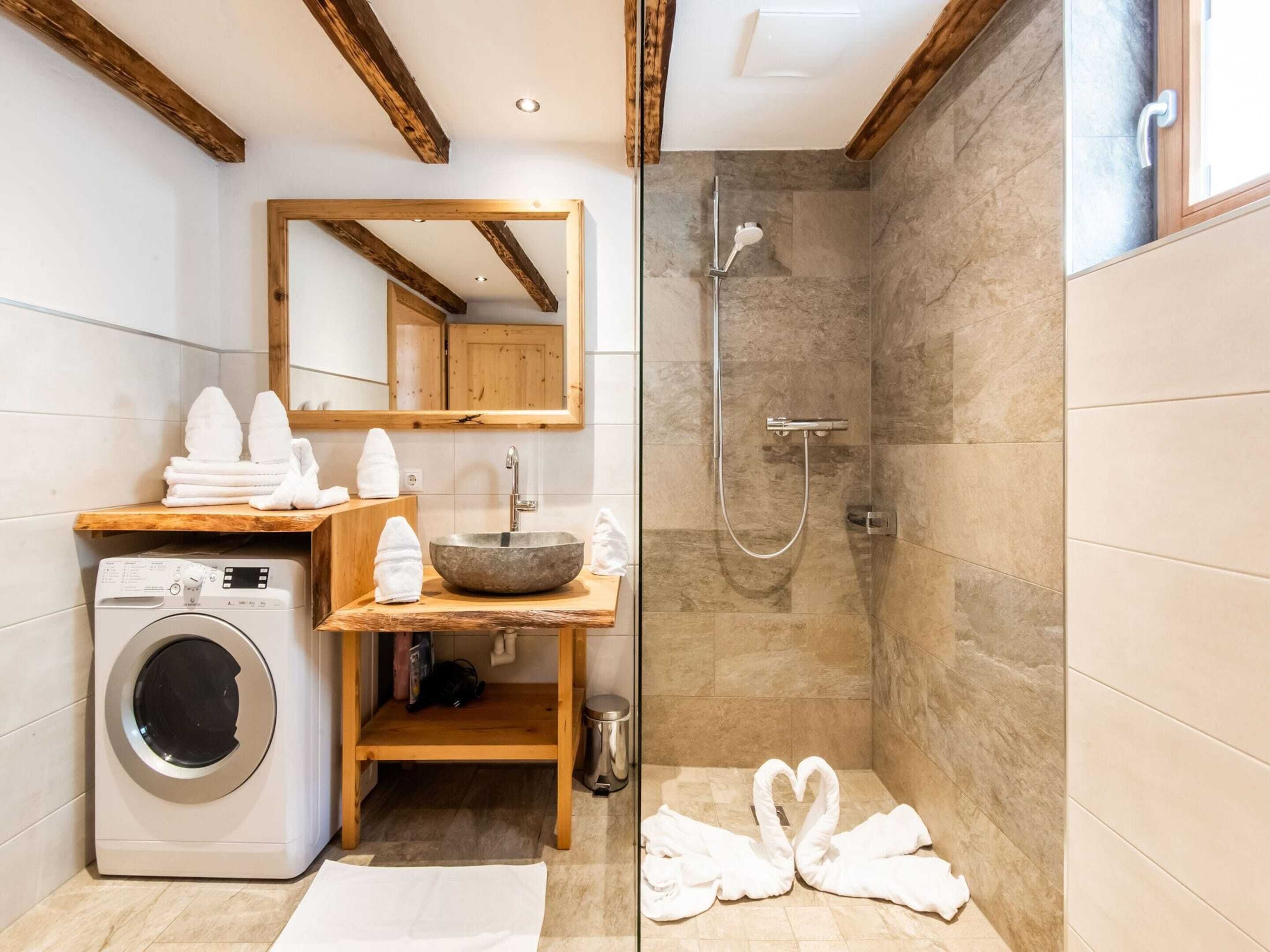 A modern bathroom with a built-in washing machine, a wooden vanity, a large mirror, a shower with a glass door, and two towels shaped like swans on the floor.
