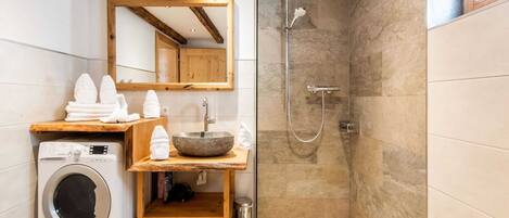 A modern bathroom with a built-in washing machine, a wooden vanity, a large mirror, a shower with a glass door, and two towels shaped like swans on the floor.