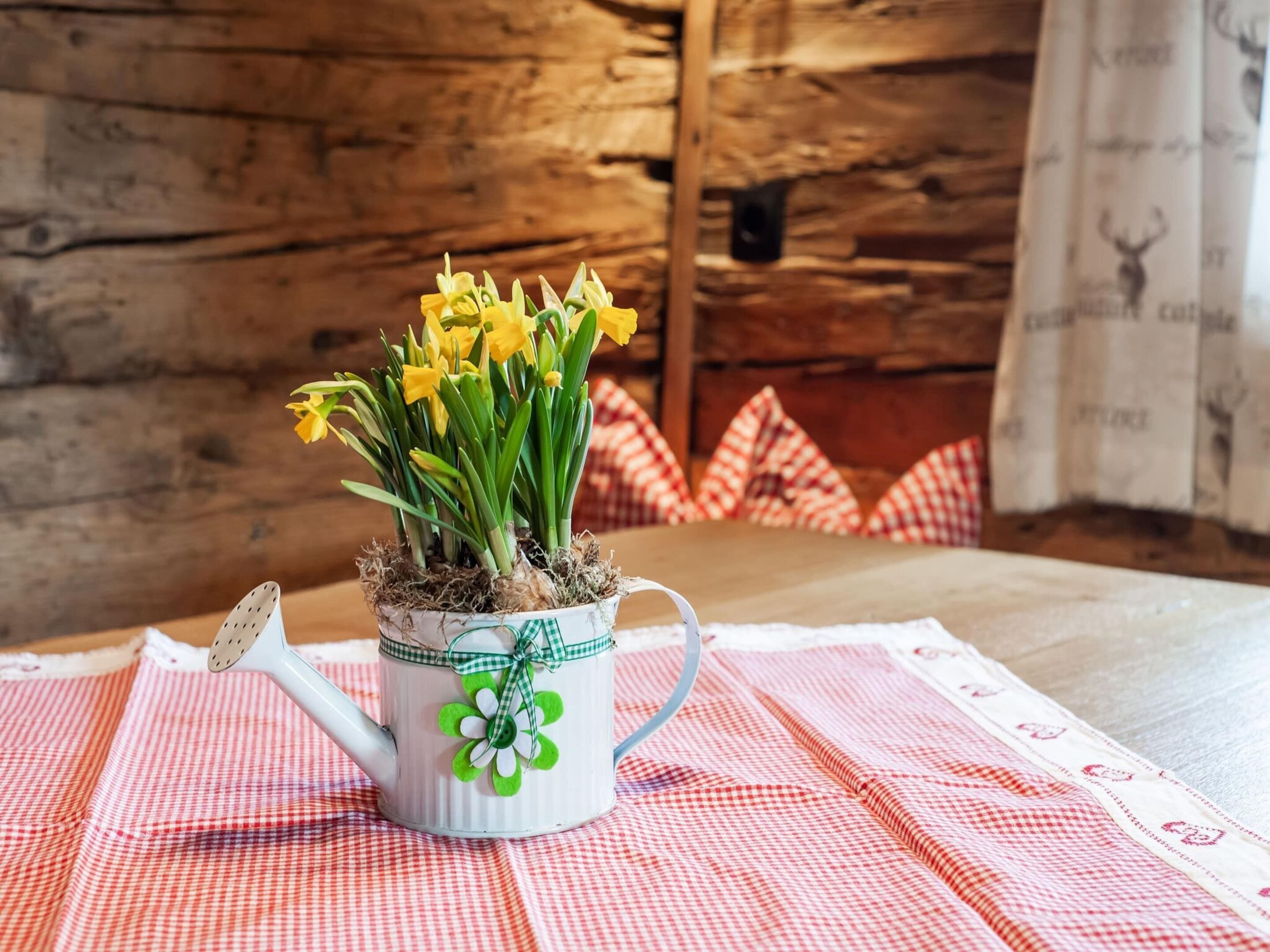 A white ceramic watering can with a green and white floral design, filled with yellow daffodils, placed on a red and white checkered tablecloth.