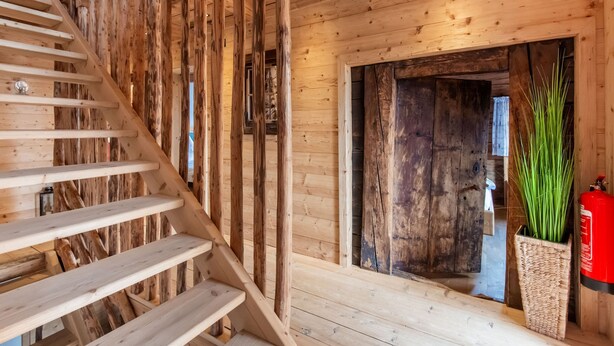 A wooden staircase with a handrail leading to a room with a large wooden door and a potted plant. - Spacious Holiday Home With Sauna (Fuegenberg)