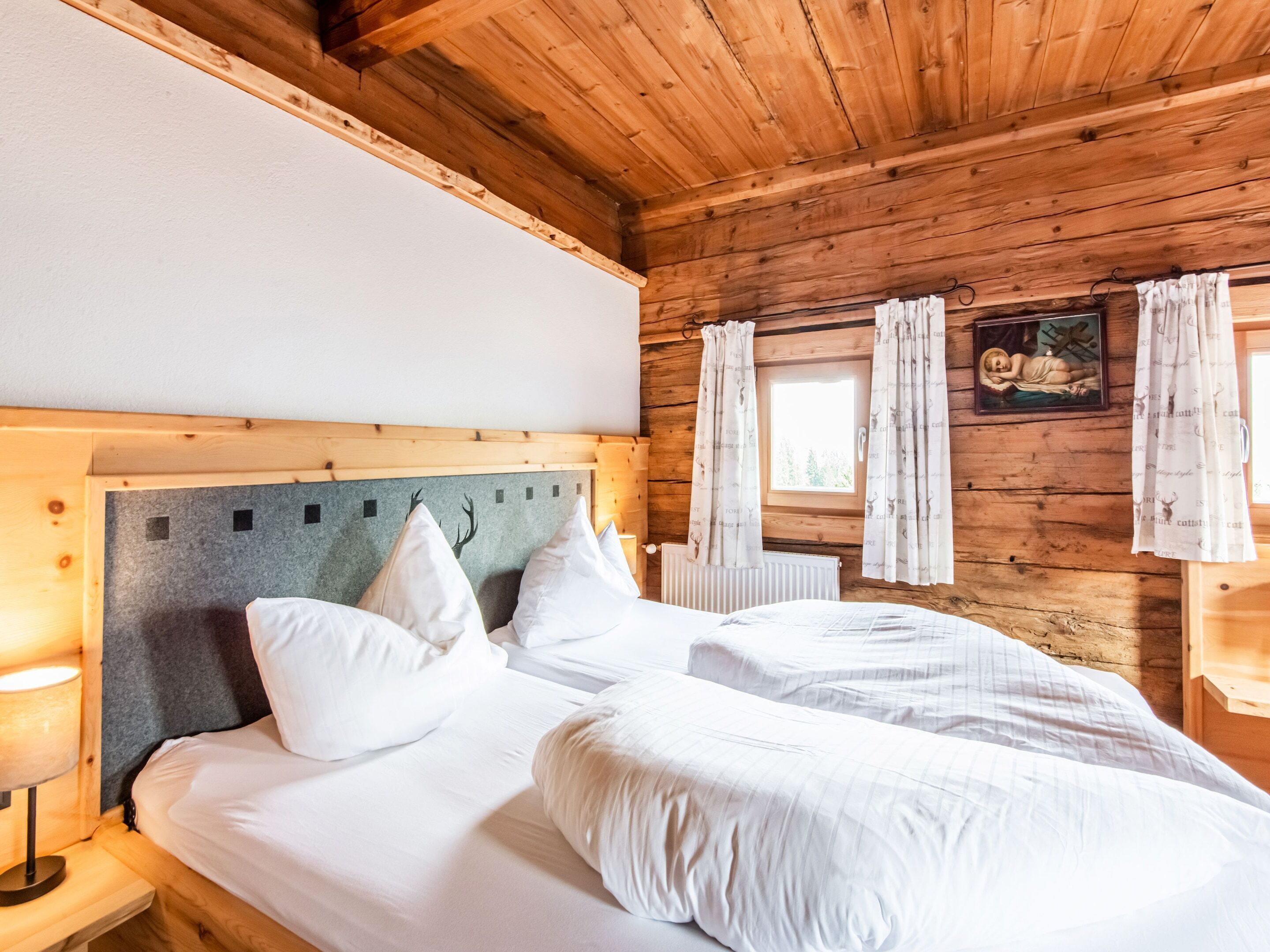 A wooden cabin bedroom with two beds, a headboard with a deer motif, and a window with curtains.