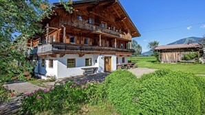 A traditional wooden chalet with a balcony, surrounded by lush greenery and a clear blue sky.
