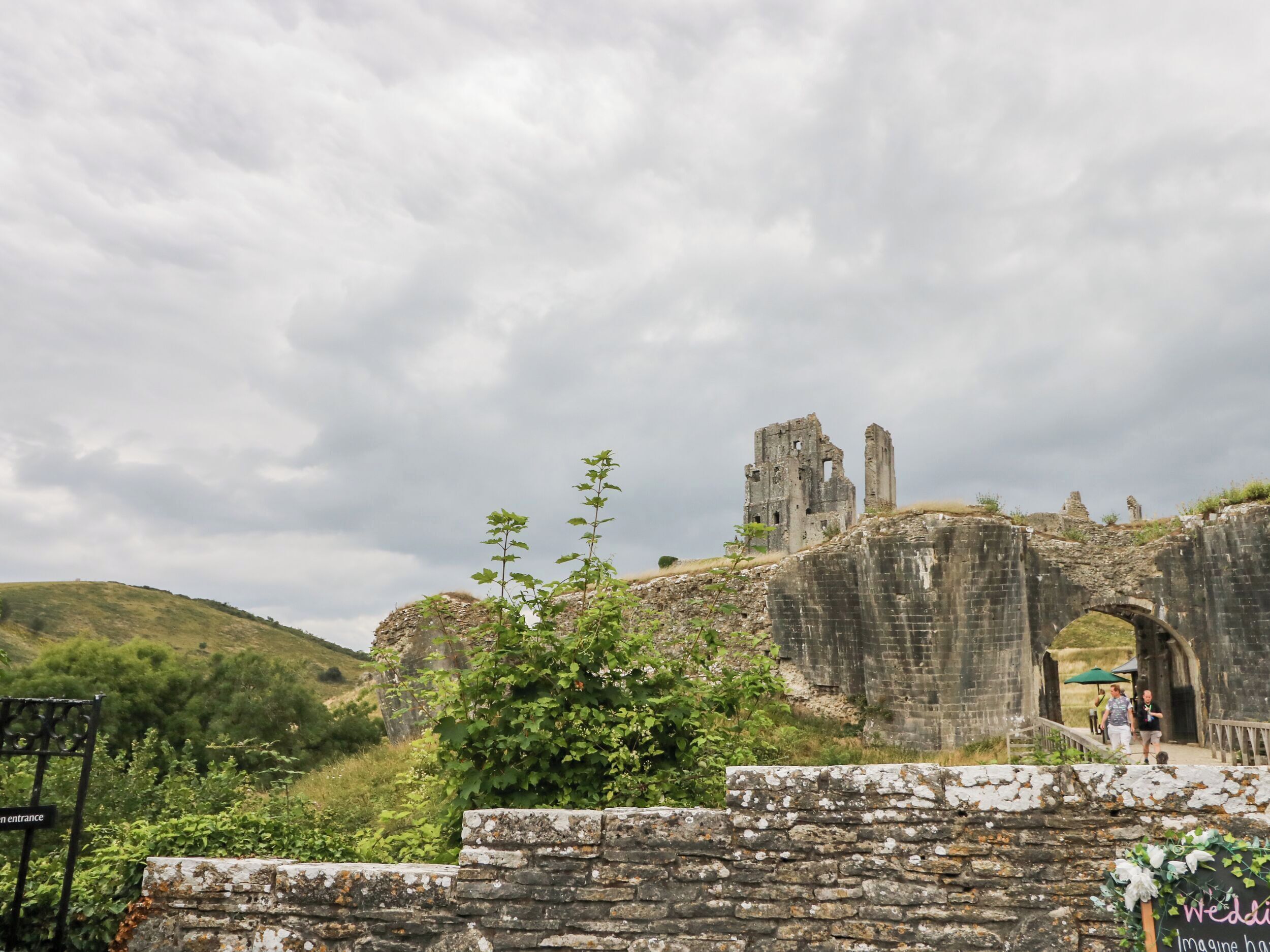 A historic stone bridge with ruins, a sign indicating an entrance, and a green umbrella in the background.