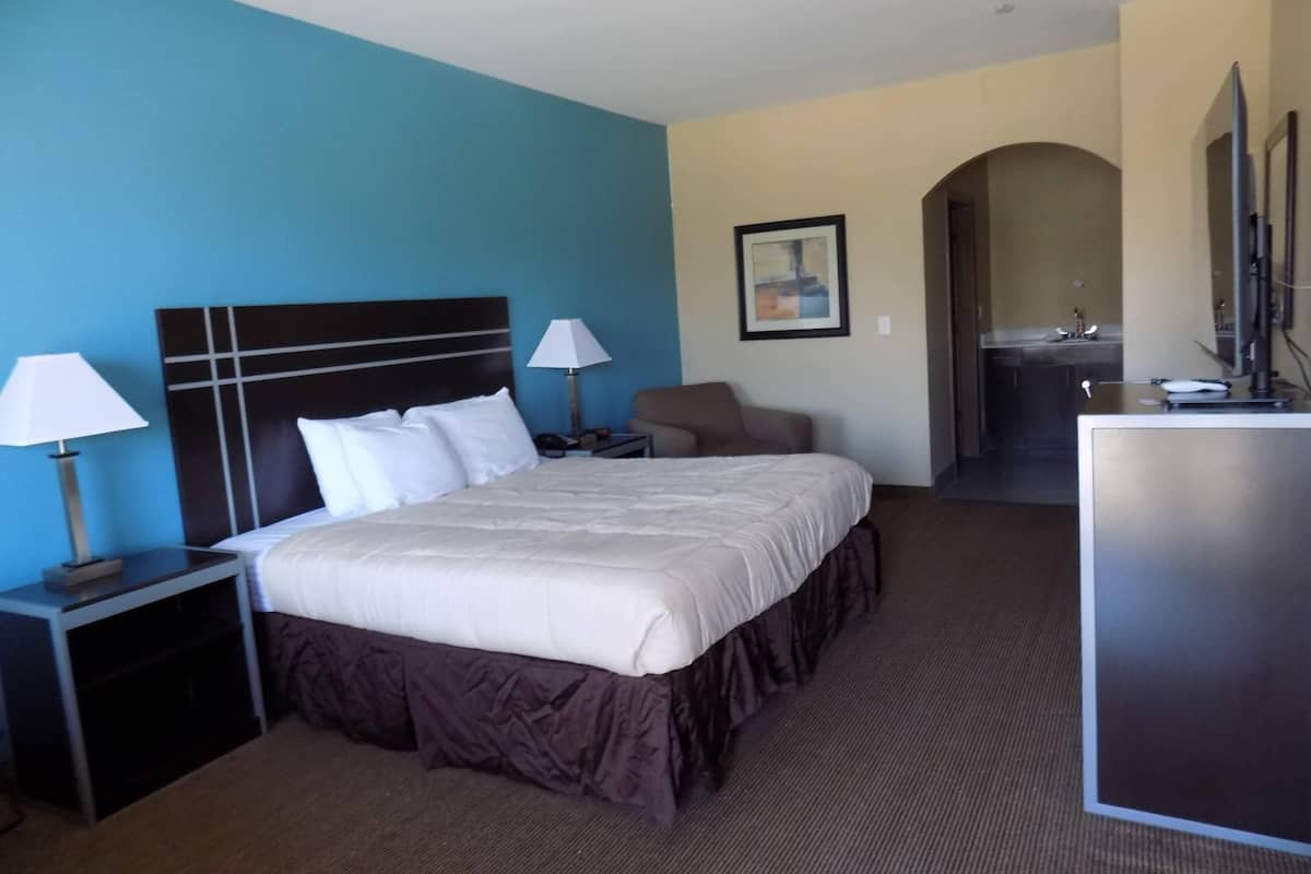 A hotel room with a large bed, two bedside tables with lamps, a chair, a desk with a mirror, and a bathroom visible through an archway.