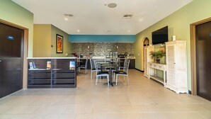 Een hotel lobby met een receptiebalie, een eetgedeelte met stoelen en een tafel, een kitchenette en een aan de muur bevestigde televisie.